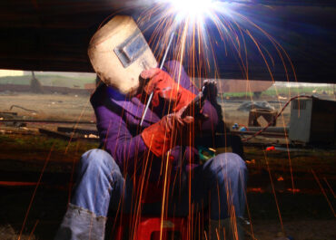A welder in helmet and red gloves is working on the underside of project while sitting on a stool